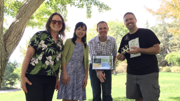Four researchers pose with a sign in a botanical garden featuring a compound model and description, smiling in the shade of a tree on a sunny day.
