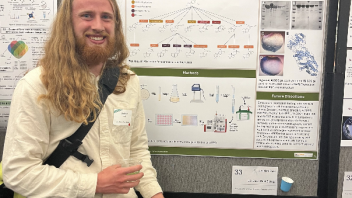 A student presenting a poster on terpene synthase research at UC Davis smiles while holding a plate of fruit and snacks at a science symposium.