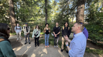 A group of students listens to a researcher speaking outdoors on a shaded forest trail, likely during a field lecture or guided educational walk.